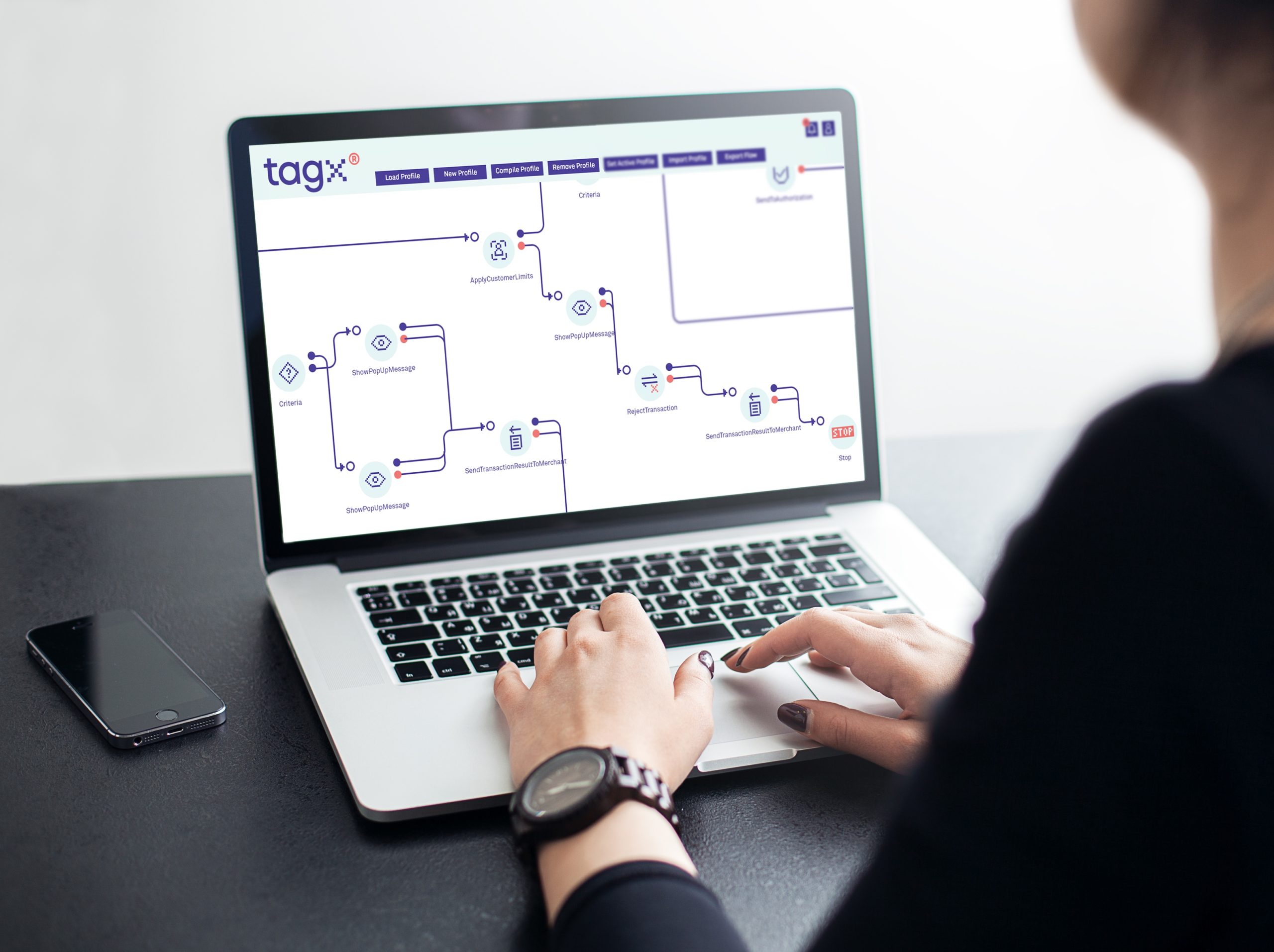 A person typing on a laptop displaying the TAGx workflow interface, showing a visual flowchart of processes and criteria. The interface features options like 'Load Profile,' 'New Profile,' and 'SendTransactionRequestToMerchant,' with a smartphone placed next to the laptop.